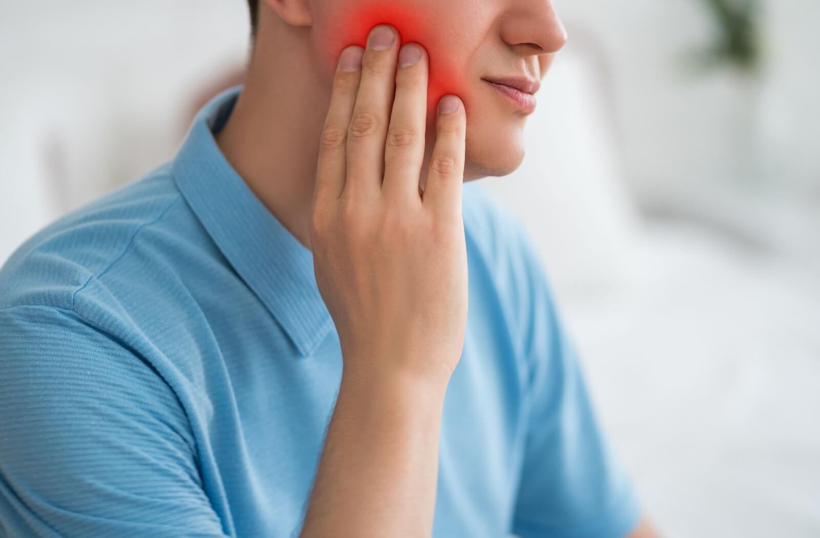 Man in a blue shirt holding his cheek with redness, indicating jaw or tooth discomfort.