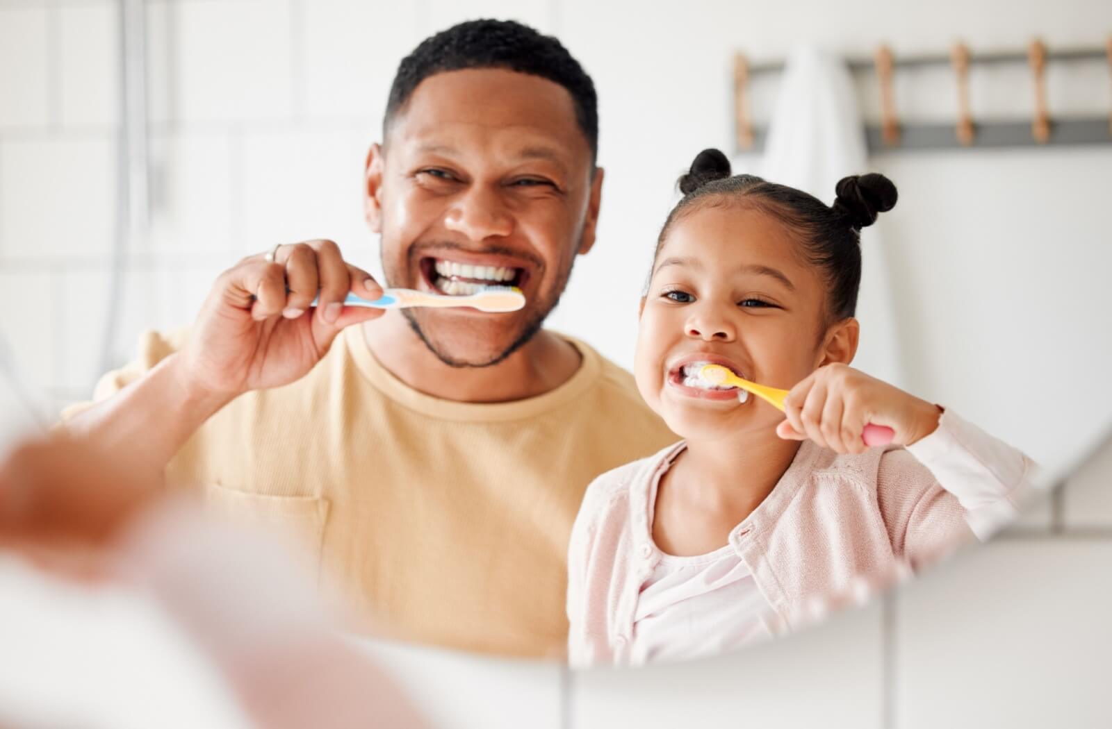 A father and daughter smile as they brush their teeth in front of a mirror.