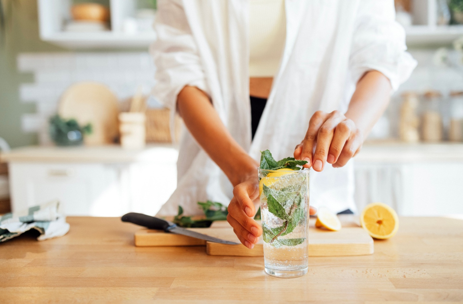 A person standing in a brightly lit kitchen adding mint and freshly cut lemon slices to a glass of iced seltzer water.