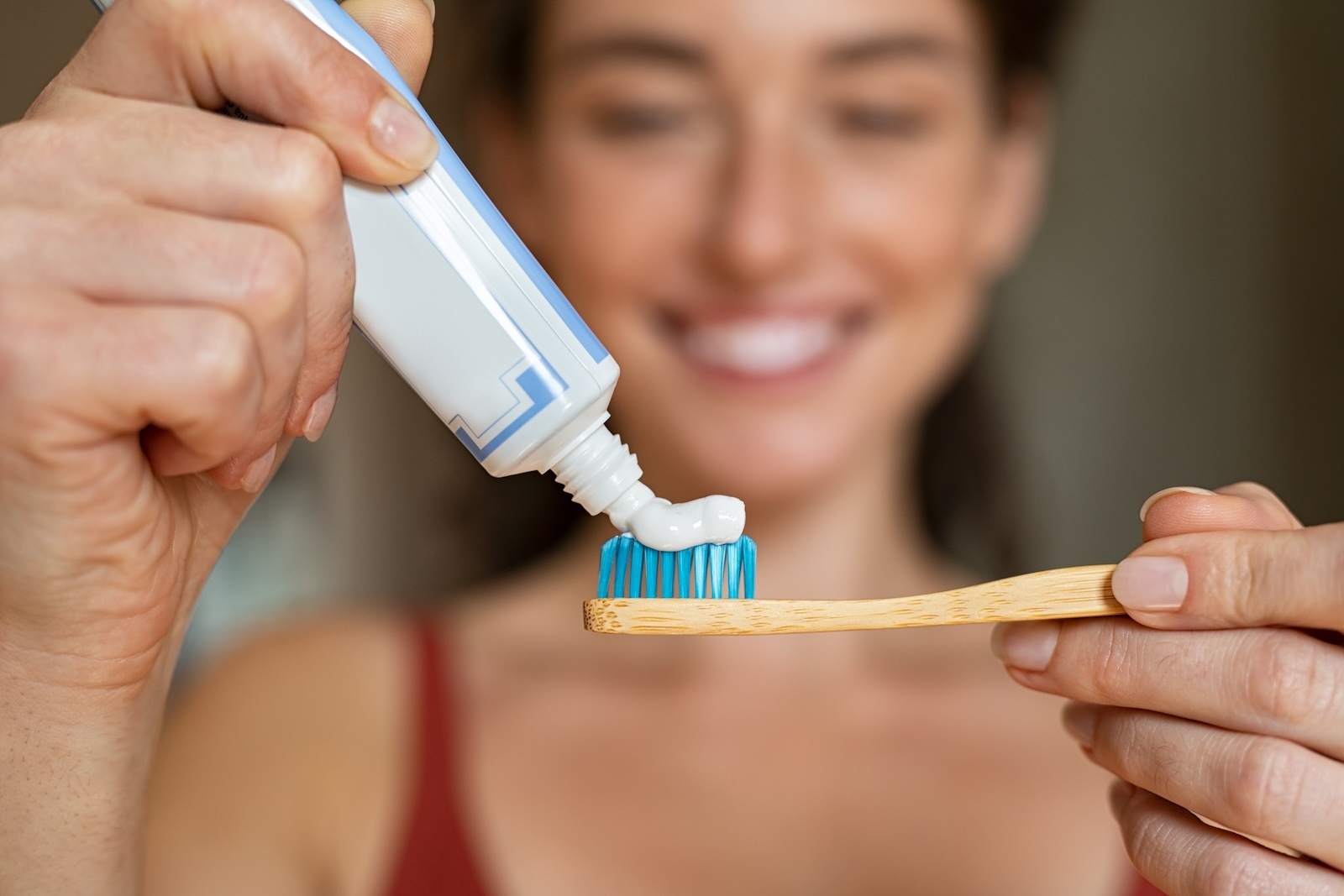 Woman in blurred background holding a wooden toothbrush in the foreground with blue bristles and squirting white toothpaste.