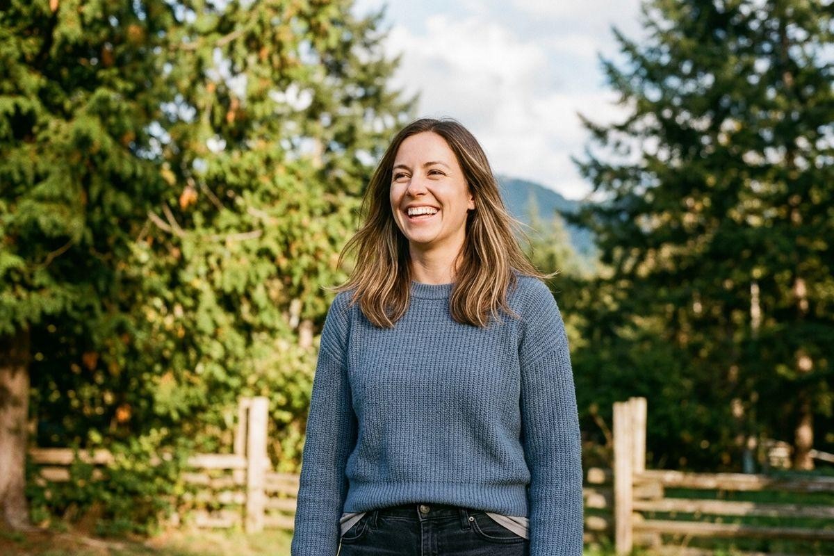 A smiling adult in a blue sweater stands outdoors by a wooden fence and green trees, looking off-camera.